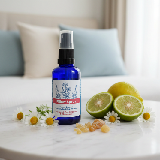 A cobalt blue glass bottle of Pillow Spray with a black spray top, set on a white marble table with sliced limes, chamomile flowers, and honey crystals. The background shows a softly blurred bedroom setting with light blue pillows.