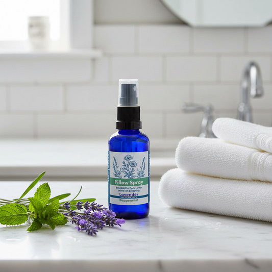 A cobalt blue spray bottle of Peppermint Pillow Spray stands on a white marble countertop in a bright bathroom setting, flanked by fresh mint and lavender sprigs on the left and a stack of folded white towels on the right.