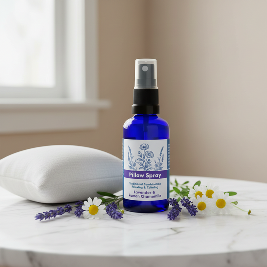 A cobalt blue glass spray bottle labeled Pillow Spray stands on a white marble surface beside a white pillow, lavender sprigs, and small white flowers in a softly lit indoor setting.