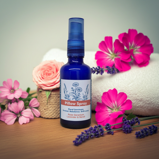 A cobalt blue glass spray bottle labeled 'Pillow Spray' with botanical illustrations, styled on a wooden surface with pink flowers, lavender sprigs, and a rolled white towel in the background.