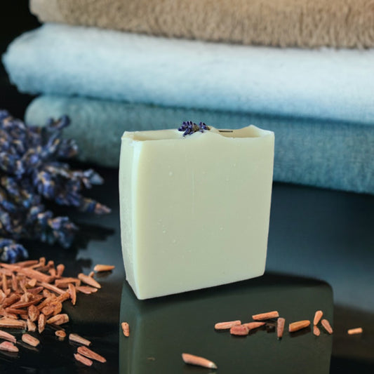 A rectangular bar of pale cream soap stands on a glossy black surface, with dried lavender sprigs and wood shavings around it and folded towels blurred in the background. Handmade lavender and cedarwood soap bar on a marble surface by Megan & Willow